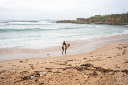 Anonymous surfer carrying surfboard gazing at sea waves rushing towards the shore. Unknown person is ready for surfing session while standing on sandy beach, engaging in extreme water sport.の写真素材