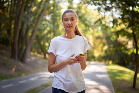 Portrait of confident female jogger listening to music through earphones while holding smartphone. Beautiful and fit woman runner in white shirt looking at camera, standing on road in park.の写真素材