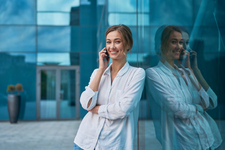 Portrait of smiling young businesswoman in white shirt talking on smartphone while leaning on glass office building. Confident female entrepreneur discussing on cellphone outside.の写真素材