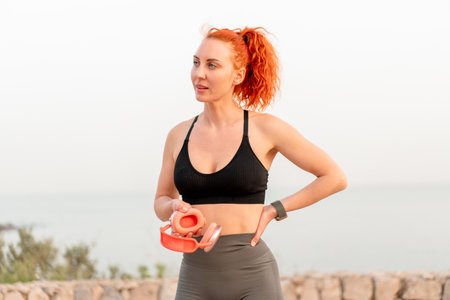 Sporty woman with red hair holding wireless headphones taking a break after tiring morning jogging. Fit female jogger in sportswear looking away. Health and fitness concepts.の写真素材