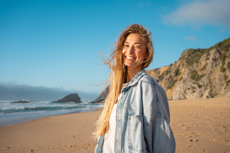 Portrait of smiling young woman with long blond hair on sandy beach. Happy female in denim shirt with sea and mountain in background. Girl enjoying vacation against blue sky.の写真素材