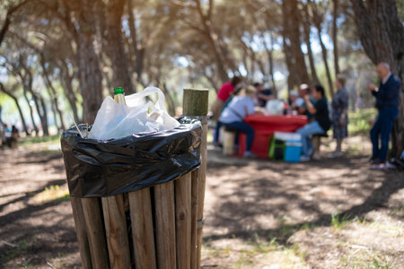 Overfilled wooden garbage bin in summer park, crowded with bottles and bags, while family and friends in the background appear blurred.の写真素材