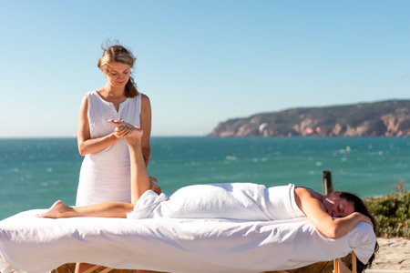 Blonde female masseur giving legs massage client lying on massage table outdoor. Spa massage on ocean shore on sunny day. Body care during summer at sea vacationの写真素材