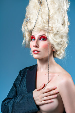Funky woman with vintage wig covering breast and looking at camera. Portrait of funny princess with red makeup wearing elegant jacket against blue background.の写真素材