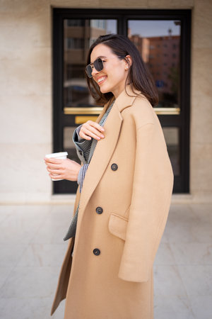Happy caucasian girl walking with cup of coffee in city on sunny day. Brunette woman wears stylish jacket coat and sunglasses. Positive emotions lifestyle conceptの写真素材
