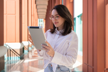 Businesswoman in glasses using digital tablet, standing outdoor dressed white shirt. Female freelancer holding tablet while walk outsideの写真素材