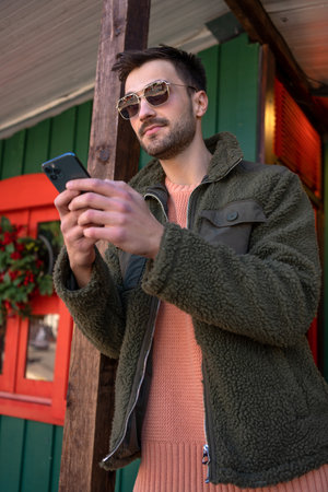 Male adult wearing sunglasses and green jacket using smartphone for sending message. Man in fashionable eyewear texting on cellphone. Modern lifestyle, connection concept.の写真素材
