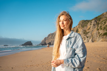Blond woman in denim shirt looking away while standing on sandy beach. Thoughtful lady with long hair near sea and mountain in background enjoying summer vacation.の写真素材