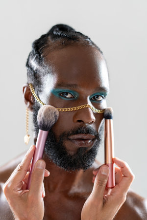African American gay man applying makeup with brushes held by crop artist. Closeup of homosexual man wearing bright eyeshadow and gold chain looking at camera against light background.の写真素材