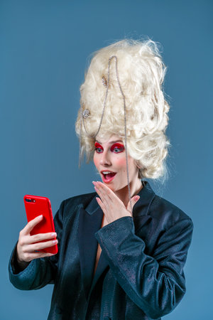 Funny woman wearing vintage wig with red makeup using smartphone for online shopping, browsing internet and networking in social media.の写真素材