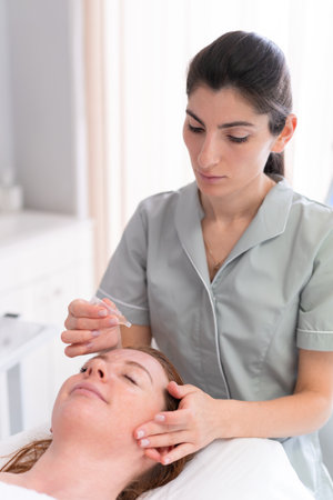 Face skin care. Beautiful woman receiving serum oil treatment in beauty salon. Cosmetologist applying cosmetic serum on head of customer at center.の写真素材