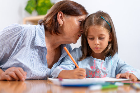 Loving grandmother whispers secret into granddaughters ear as she colors in book at home. The picture captures warm, loving moment filled with affection and artistic activityの写真素材