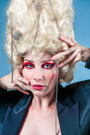 Closeup of glamourous female model wearing huge blond wig with accessories over blue background. Attractive woman with face makeup is posing in studio.の写真素材