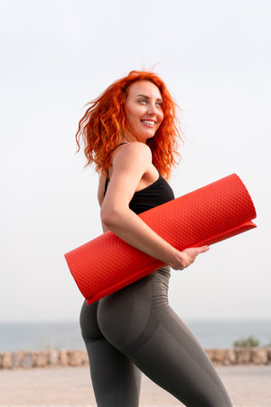 Happy jogger woman holding rolled up exercise mat against clear sky. Side view of fit young female in activewear is looking away during daytime.の写真素材
