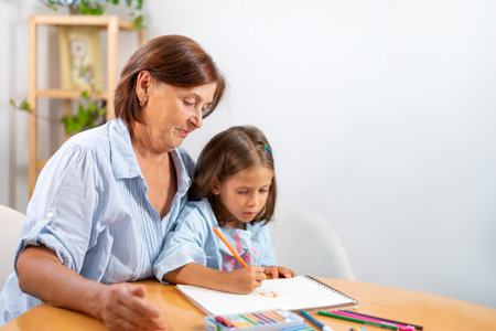 Smiling grandmother watching girl as she draws with colored pencil in book at home. The scene captures moment of admiration and encouragement as child is busy in her creative activityの写真素材