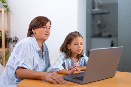 Focused granddaughter and grandmother surfing on laptop together at home. Senior woman and girl are learning from the computer during leisure timeの写真素材