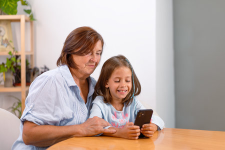 Happy granddaughter using smartphone with grandmother on table at home. Their smiling faces are full of content as they share and learn from the deviceの写真素材