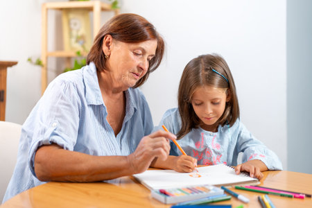 Focused grandmother and granddaughter coloring in book together on table at home. The scene is filled with creativity and warmth, as they bond over their artistic activityの写真素材