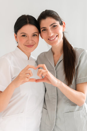 Happy female doctors making heart shape with hands at clinic. Smiling medical professionals are looking at camera while standing together in hospital.の写真素材