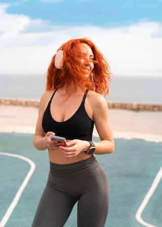 Happy sporty woman holding smartphone while standing at outdoor gym. Fit young female in sportswear is standing against sea and sky during sunny day.の写真素材