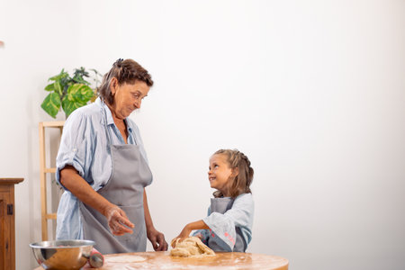 Happy girl looking at grandmother while kneading pastry dough on table at home. Little female child is learning baking from elderly womanの写真素材