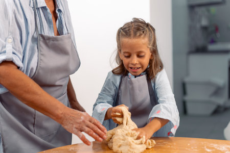 Cute girl kneading pastry dough in assistance of anonymous grandmother on table at home. Little female child is learning from elderly woman, baking conceptの写真素材