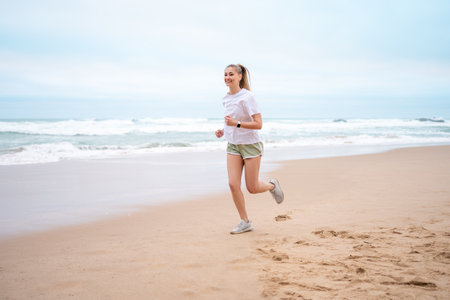 Happy sporty woman jogging on wet seashore against sky at beach. Full body of smiling female is looking away while exercising during daytime.の写真素材