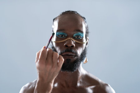 Confident African American gay model applying eye makeup over gray background. Portrait of serious...