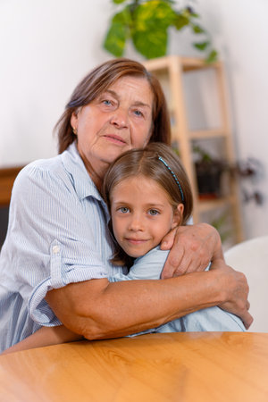 Loving grandmother and granddaughter share warm embrace at home, their faces filled with joy as they look at camera, capturing heartfelt moment of connection across generationsの写真素材