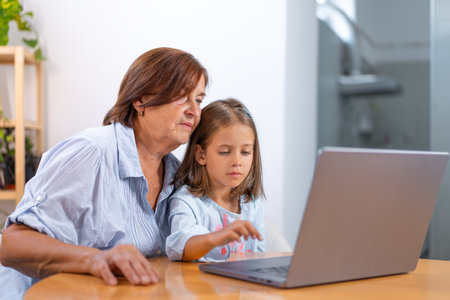 Granddaughter and grandmother sit together at table using laptop, sharing moment of learning and connection in comfort of their homeの写真素材