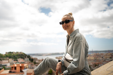 Happy female tourist sitting on retaining wall at Senhora do Monte in Lisbon viewpoint against cloudy sky. Portrait of smiling attractive woman in casualwear and sunglasses is on vacation.の写真素材