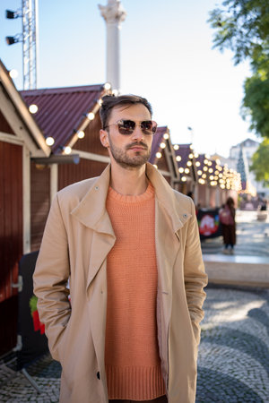 Fashionable man wearing sunglasses standing in front of decorated house. Casual good looking male posing near wooden house with festive lightbulbs. Brunette bearded male next to Christmas decorationsの写真素材