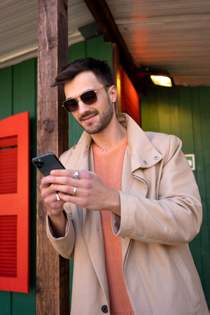 Smiling man in beige trench coat using smartphone outside green house. Handsome young male is leaning on wooden pole during daytime.の写真素材