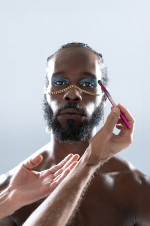 Crop hands of makeup artist applying eye makeup on African American gay model over gray background. Portrait of serious homosexual man with chain on nose is in studio.の写真素材
