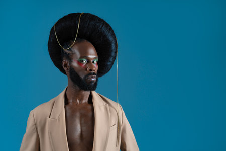 Serious African American gay model wearing huge wig with chain over blue background with mock up space. Black man in blazer is looking at camera at studio.の写真素材