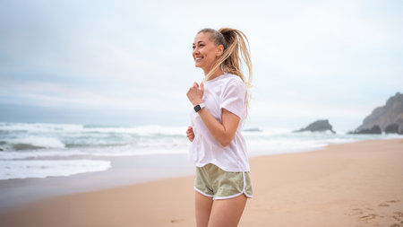 Smiling sporty woman jogging on seashore against sky at beach. Happy female is looking away while...