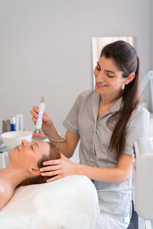 Happy cosmetologist applying iontophoresis on clients face. Pretty Caucasian lady getting physiotherapeutic treatment for her facial skinの写真素材