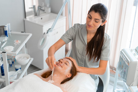 Professional cosmetology: Smiling massage therapist applying face mask to woman with brush at salon. From above of relaxed female customer is getting pampered in spa by beauticianの写真素材