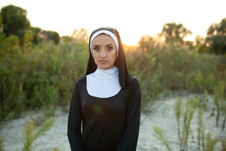 Serious nun looking at camera while standing on field during sunset. Young woman in religious clothes is surrounded by plants outdoorsの写真素材