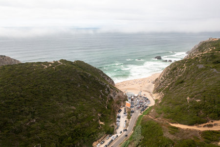 Aerial drone view of parking lot nestled between hills at Adraga Beach in Sintra, Portugal. The scenic landscape contrasts with orderly parked cars, highlighting natural beauty of coastal settingの写真素材