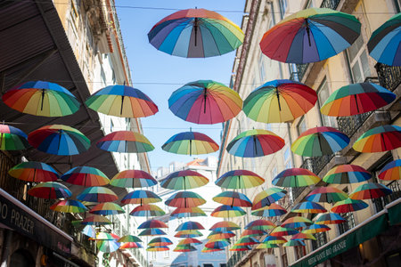From below shot of rainbow umbrellas hanging in city. Multicolored objects are decorated amidst buildings during daytimeの写真素材