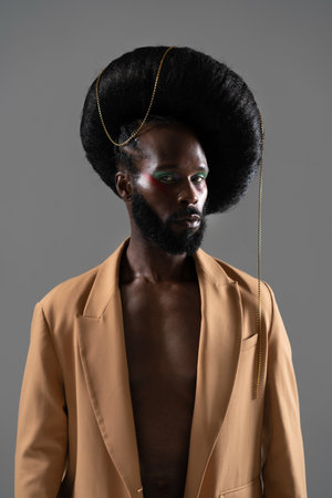 Serious African American gay model wearing huge wig with chain over gray background. Black man in brown blazer is looking at camera in studioの写真素材