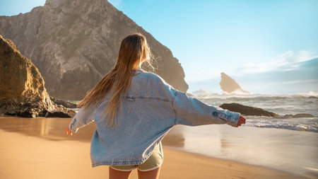 Anonymous woman in denim shirt standing with arms outstretched on seashore against blue sky at beach during summer. Back view of female tourist with long blond hair is enjoying vacationの写真素材