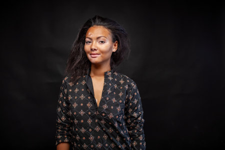 Smiling woman in traditional black kurta standing against black background. Young woman with vitiligo skin disorder looking at camera in studioの写真素材