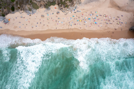 Aerial view of unspoiled ocean beach with turquoise waters and pristine sands. Tourists with parasols relax on sandy seashore, enjoying a peaceful vacationの写真素材
