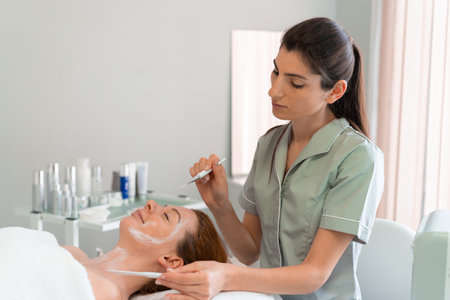 Professional masseuse in uniform applying face mask with brush on womans face at cosmetology salon. Relaxed female customer enjoying pampering spa treatment by beauticianの写真素材