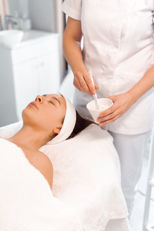 Crop dermatologist preparing face pack for female client lying on massage table at salon. Relaxed young woman undergoing facial treatment in beauty spaの写真素材