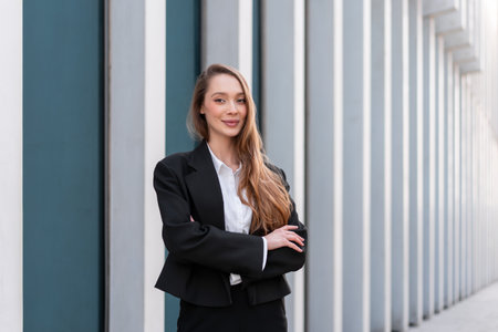 Portrait of smiling woman in business suit standing arms crossed. Young businesswoman with long hair on sidewalk along building in city. She is confidently looking at cameraの写真素材