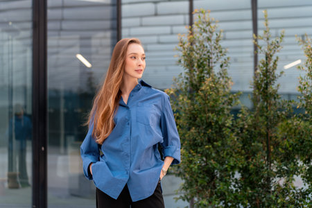 Young businesswoman in blue shirt standing in front of glass building. Beautiful woman with long...