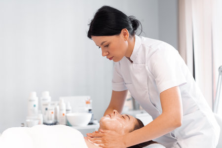Massage therapist providing facial massage to woman at cosmetology clinic. Relaxed female client is getting pampered in spa by professional beauticianの写真素材